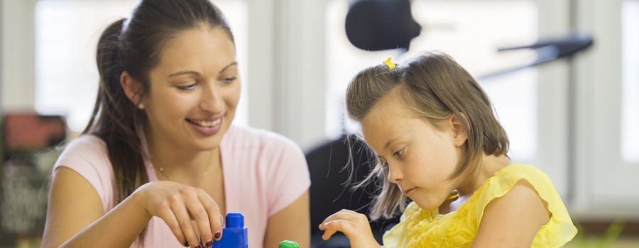 A little girl with cerebral palsy is trying to stack toy blocks with the help of her caregiver.